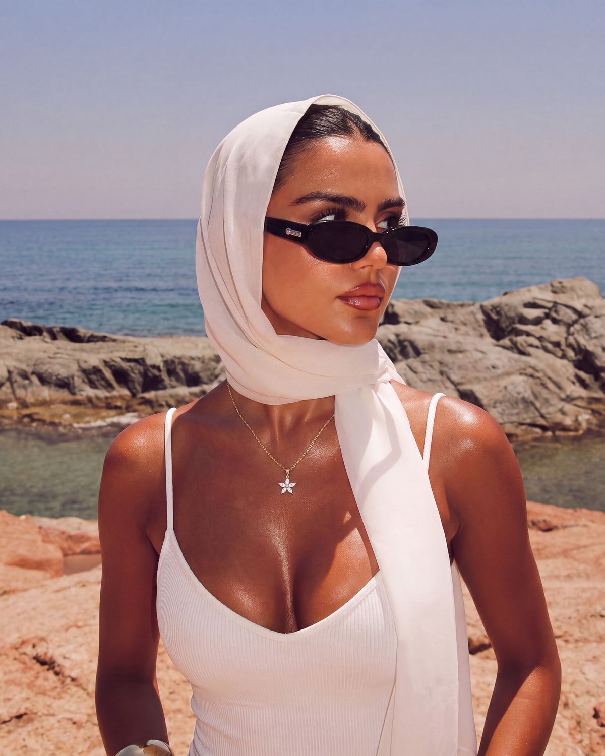 Modelo con pañuelo blanco en la cabeza y lentes de sol junto al mar con el collar Fleur de colgante de flor